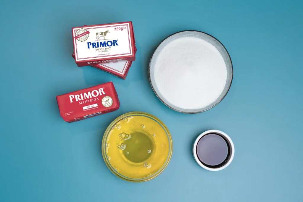 Em uma superfície azul, há dois pacotes de manteiga da marca Primor, uma tigela de açúcar, uma tigela com ovos quebrados e uma pequena tigela de extrato de baunilha.